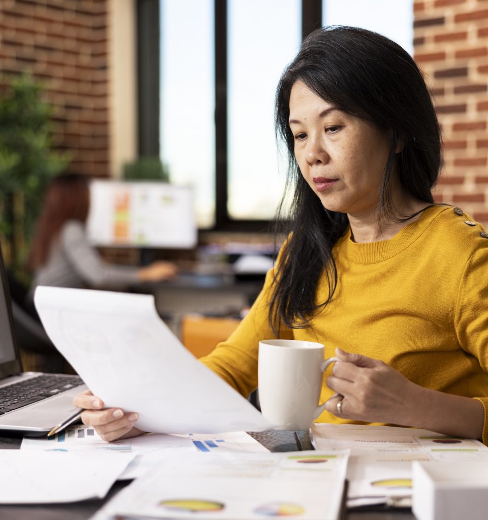 Portrait of female business advisor holding a white cup and paper, seated at office desk, busy preparing a presentation. Asian project manager with coffee in hand, reviewing her documents on table.